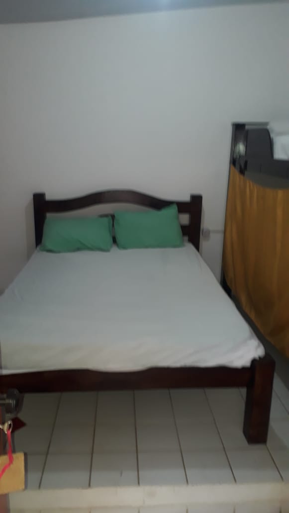 A spacious bed with light-colored bedding is positioned on a tiled platform. Two green cushions rest against the headboard. A bunk bed is visible in the background, draped with yellow fabric, contributing to the overall cozy setting.