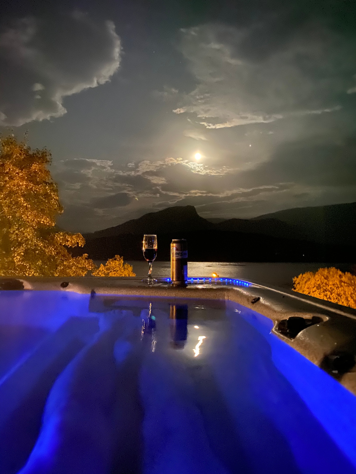 A hot tub is illuminated by soft blue lighting, with a wine glass placed on the edge beside a can. The scene features a moonlit sky and silhouettes of mountains in the distance, creating a serene atmosphere.