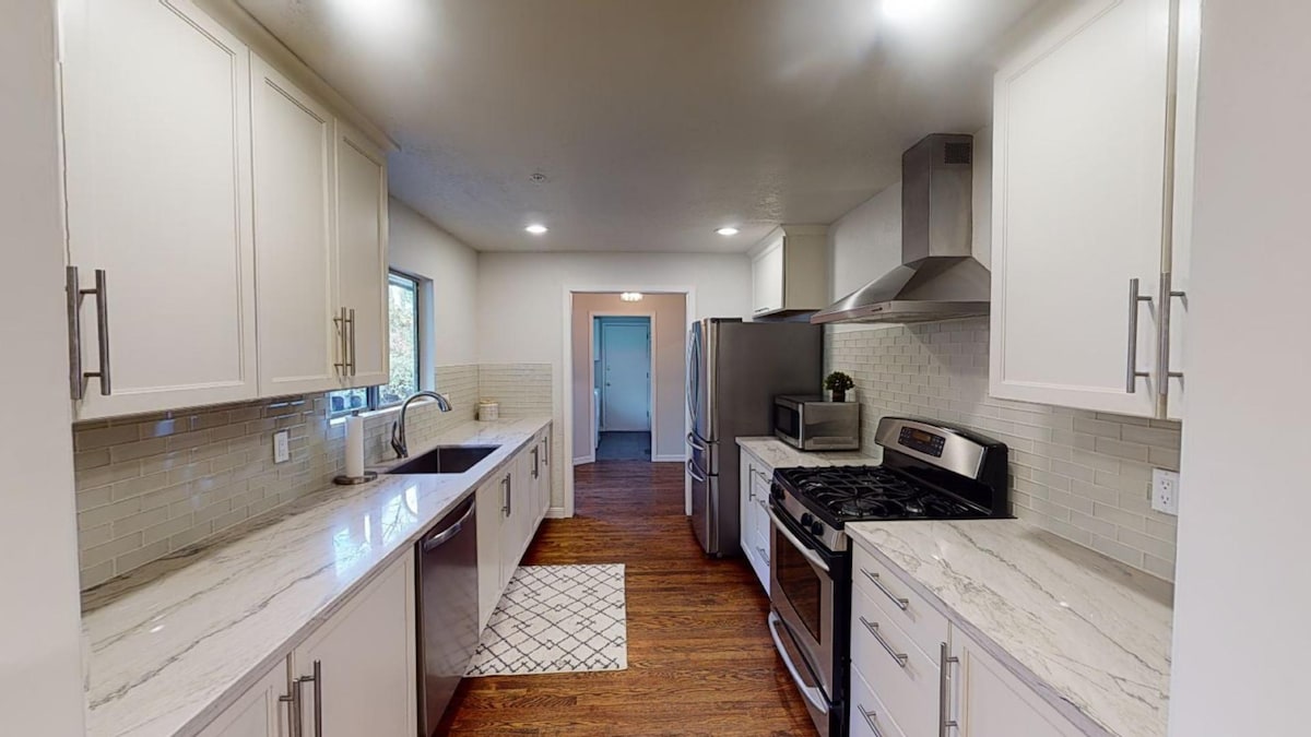 The kitchen is equipped with modern appliances, including a stainless steel refrigerator, gas stove, and microwave. White cabinetry contrasts with gray marble countertops, while a double sink is positioned under a window. Soft lighting enhances the space, which features wood flooring and a patterned tile area.