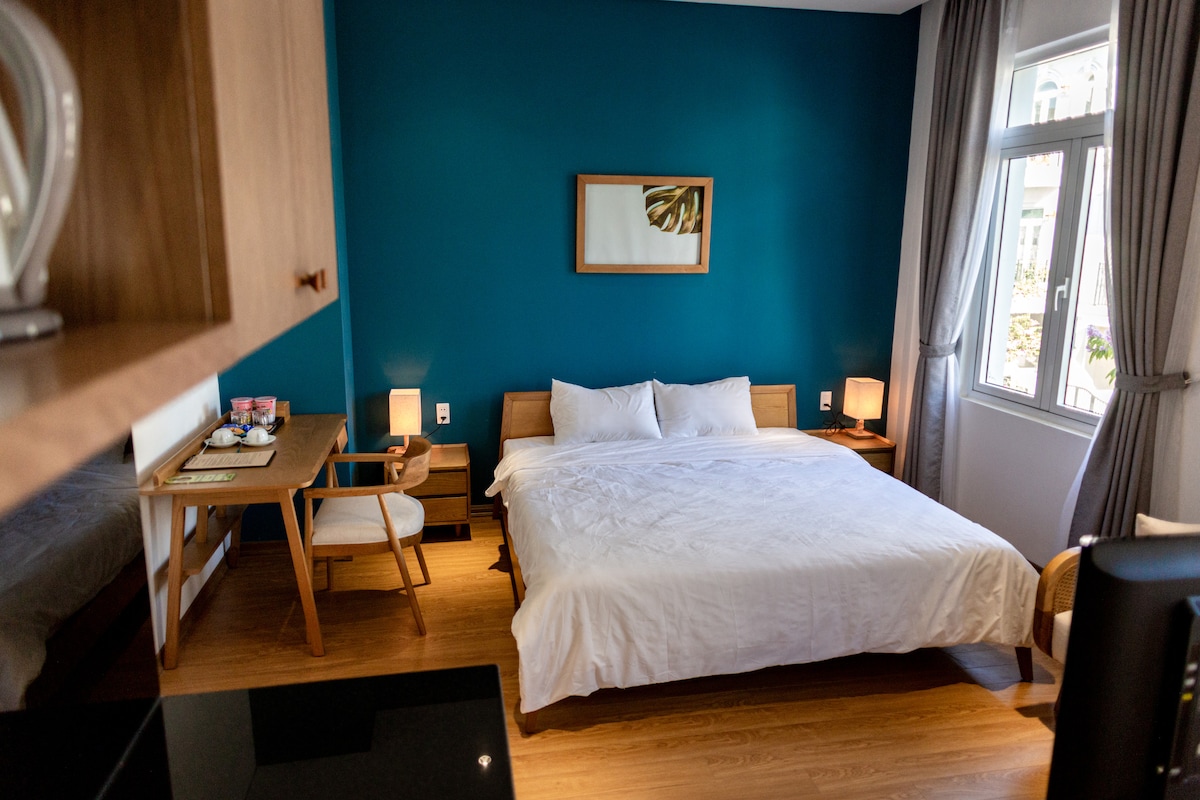 A spacious room is presented with a large bed dressed in white linens against a vibrant blue wall. Two bedside lamps provide soft lighting. A wooden desk and chair occupy one corner, with a view of the outside visible through the large windows adorned with light curtains.