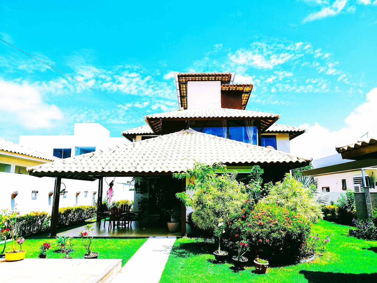 The exterior of the house displays a charming architectural style with a tiled roof and multiple levels. Lush green gardens are visible, complemented by vibrant flowers and well-maintained lawns. A spacious terrace area provides shaded seating, contributing to the inviting outdoor ambiance.