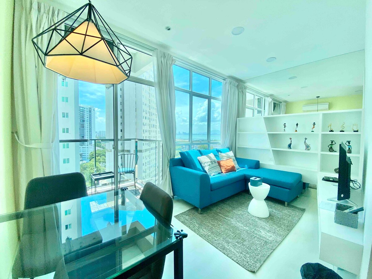 A bright living area is illuminated by natural light through large windows. A cozy blue sofa is positioned next to a small, round coffee table. A dining table with black chairs is visible, and decorative elements are displayed on shelves. A partial sea view can be seen through the glass doors.