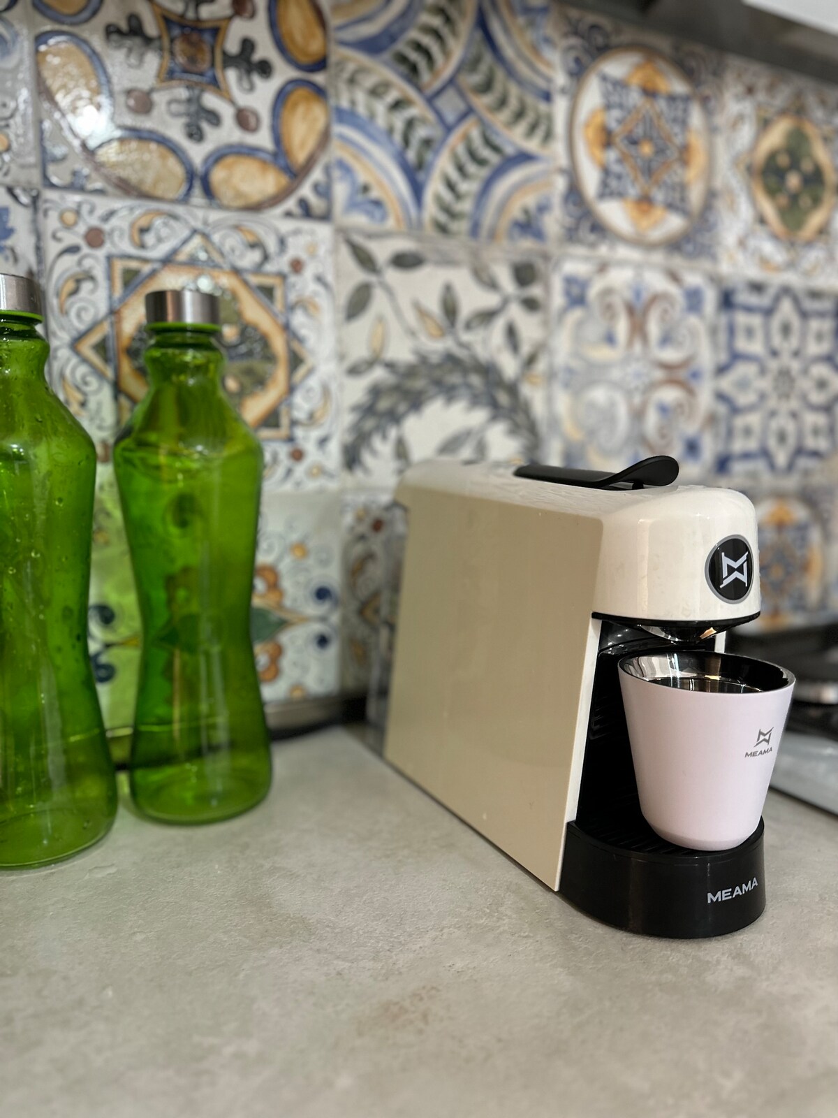 A coffee machine is positioned next to two green glass bottles on a kitchen countertop. Colorful patterned tiles provide a decorative backdrop, enhancing the kitchen's aesthetic. The coffee machine features a white and black design, with a cup placed below the spout.