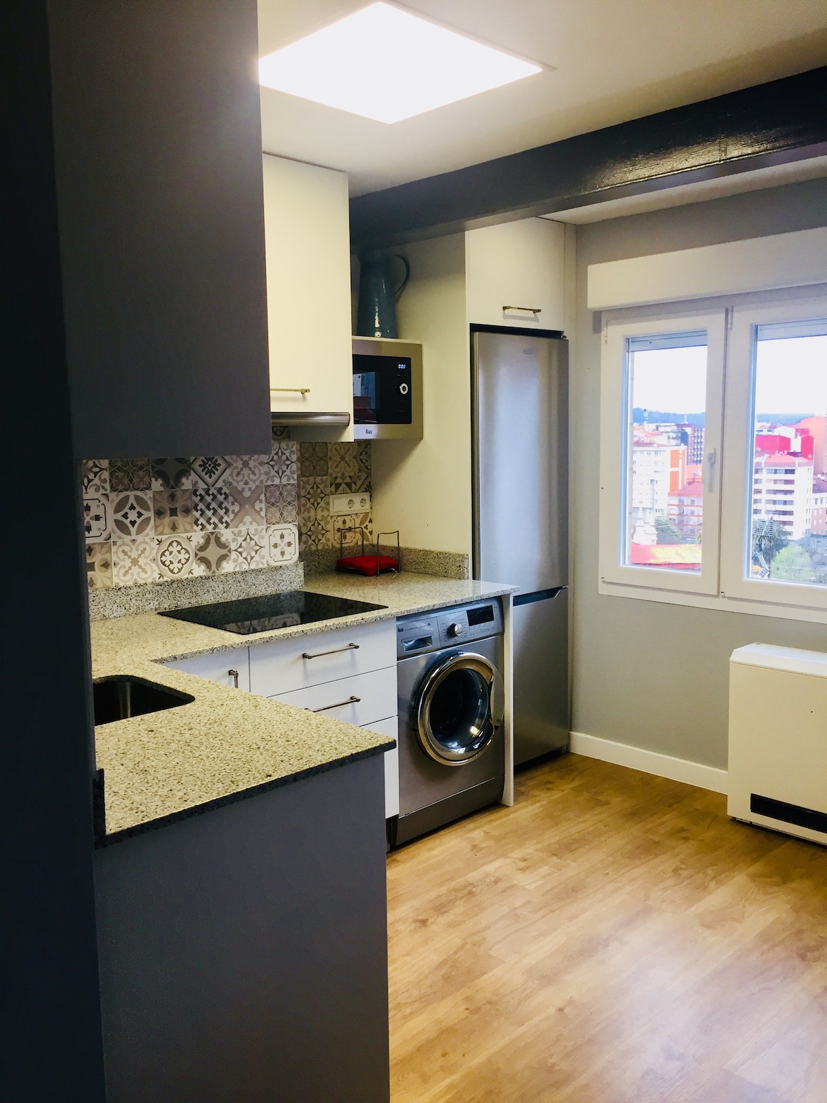 A modern kitchen is featured, equipped with a washing machine and a refrigerator. Light-colored cabinets line the walls, complemented by a textured backsplash. A window allows natural light to illuminate the space and offers views of the cityscape outside.