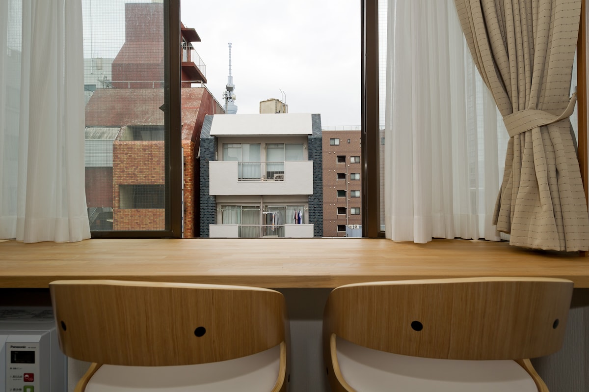 A wooden workspace is positioned in front of large windows, offering a view of nearby buildings and the Tokyo Skytree in the distance. Two simple chairs are placed at the desk, enhancing the functional design of the area.