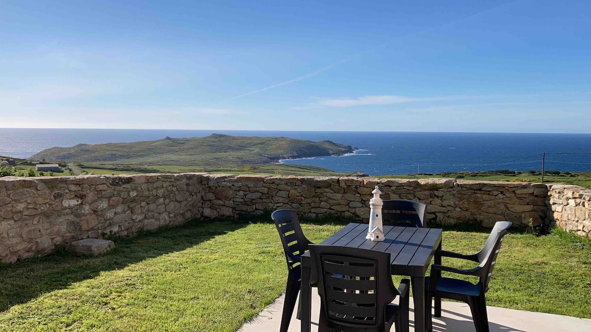 An outdoor terrace features a black dining table surrounded by four chairs, overlooking panoramic views of the Atlantic Ocean and the landscape beyond. The area is bordered by a low stone wall and includes a grassy space, ideal for enjoying meals or relaxing outdoors.