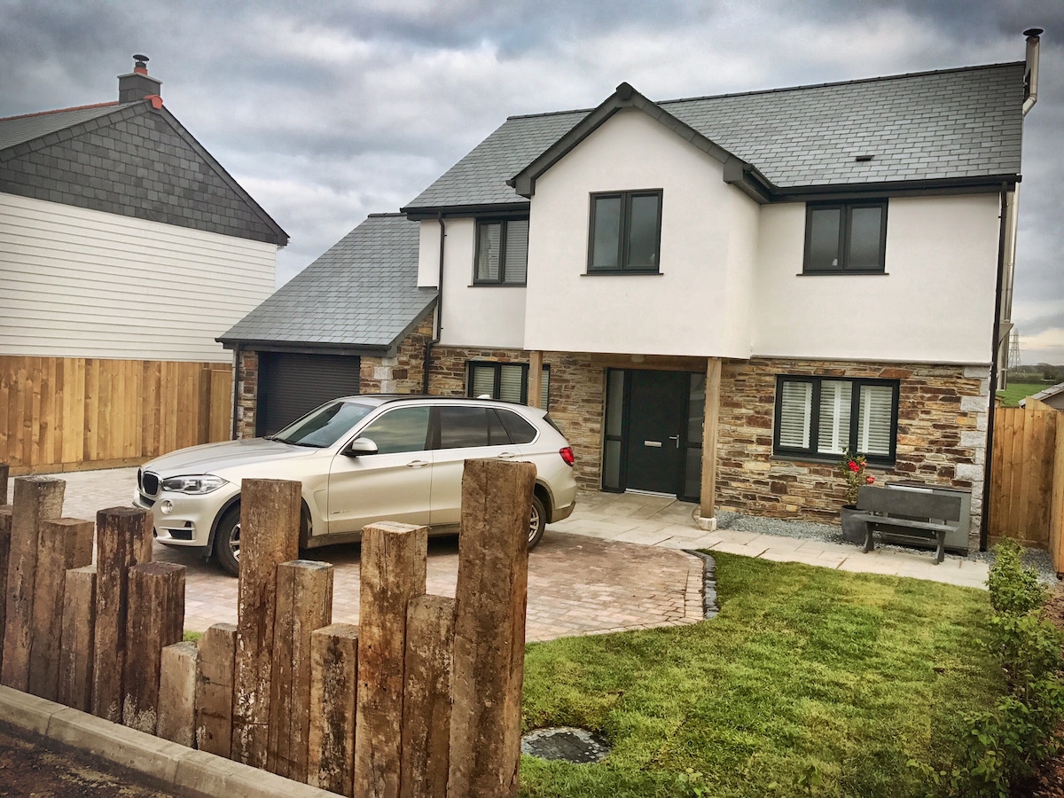 A modern two-story house features a mix of stone and white siding, with large windows on the upper level. A paved driveway accommodates a vehicle, while a manicured lawn and wooden fence provide a welcoming entry. The overall appearance reflects a contemporary architectural style.