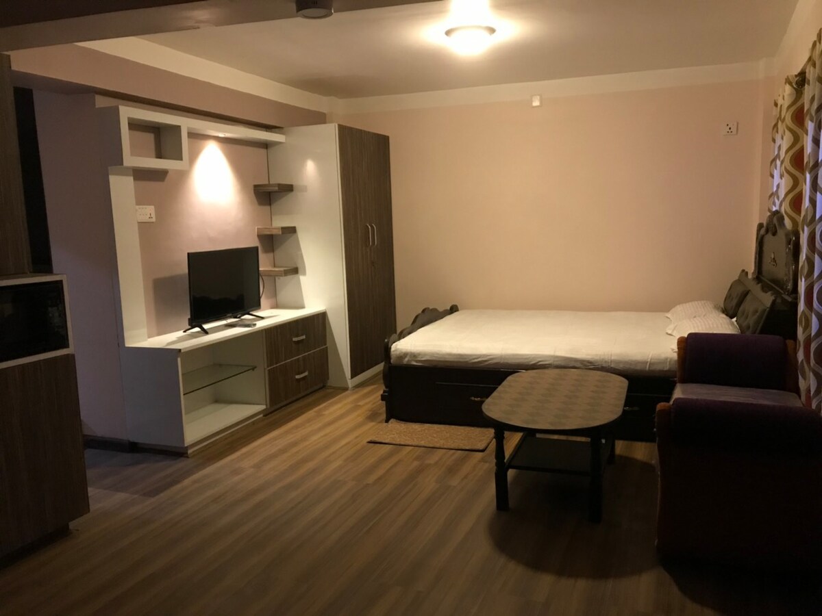 A compact living space features a spacious bed, a wall-mounted television on a modern cabinet, and a small coffee table. A comfortable chair adds to the seating options. Warm lighting enhances the cozy atmosphere, with light hardwood flooring complementing the neutral-toned walls.