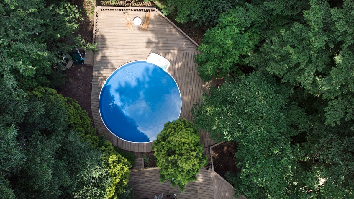 An aerial view of a circular saltwater pool is surrounded by a wooden deck, nestled within a lush landscape of dense trees. The serene blue water reflects the sky, creating a tranquil focal point amidst the greenery.