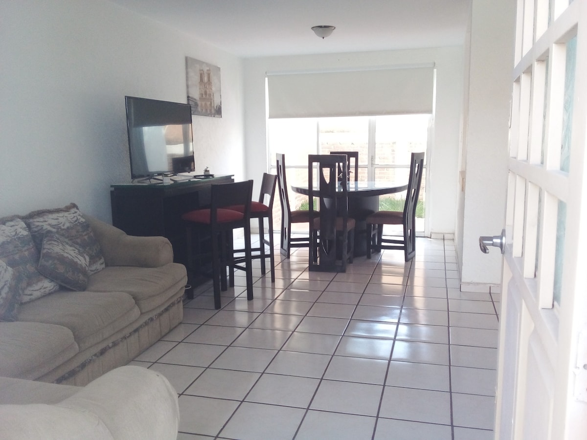 A light-filled living area features a comfortable sofa and a dining table surrounded by chairs. Large windows allow natural light to illuminate the space, while a television is positioned on a stand against one wall.