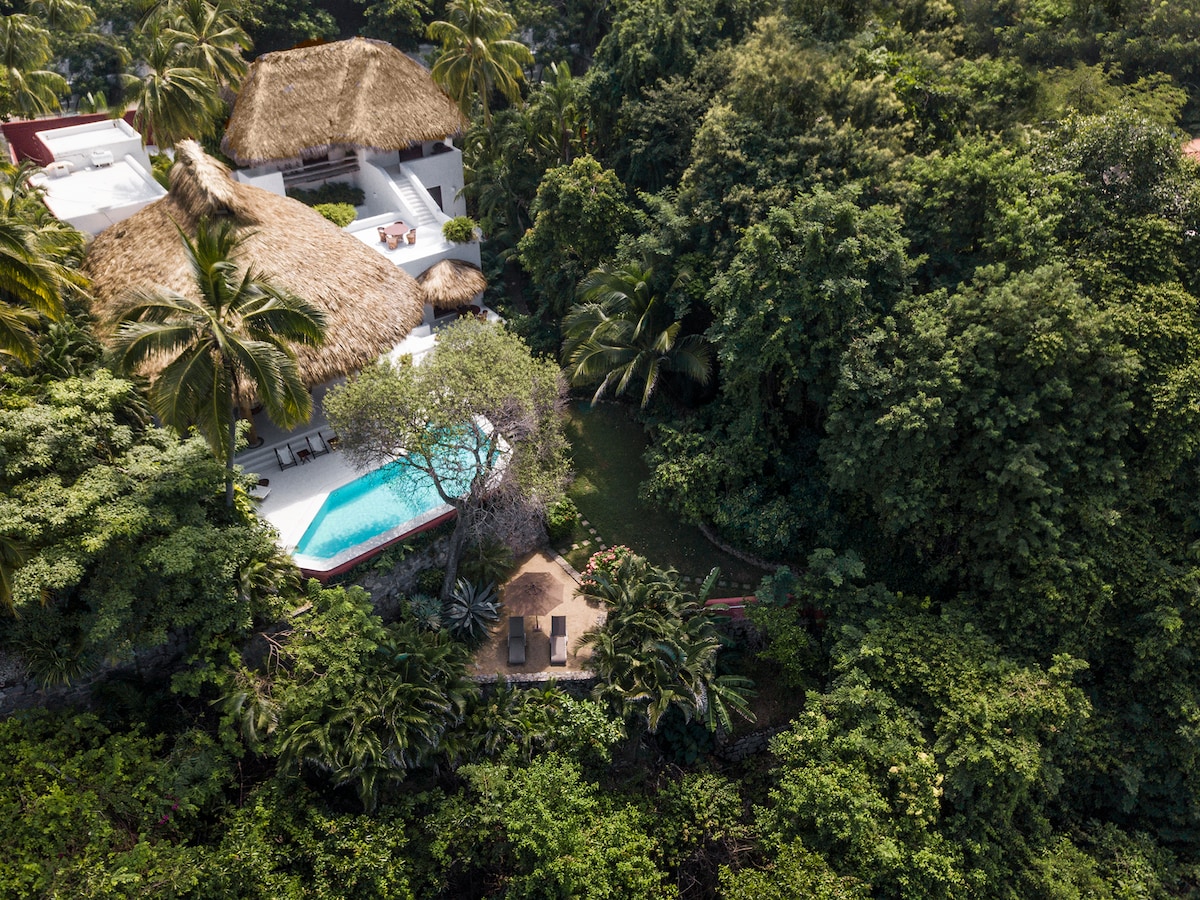 An aerial view reveals a private villa nestled among lush greenery, featuring a thatched roof and a serene swimming pool. Sun loungers are positioned near the pool, complemented by dense vegetation surrounding the property, creating a tranquil atmosphere.