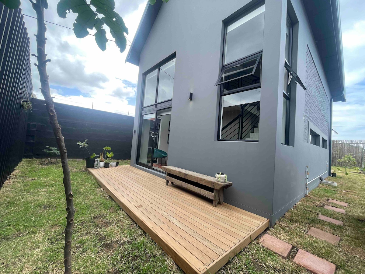 A well-maintained exterior features a wooden deck, providing an outdoor seating area. The modern house, with large windows, is complemented by a green lawn and stone pathway. Scenic clouds are visible in the sky, adding to the fresh outdoor environment.