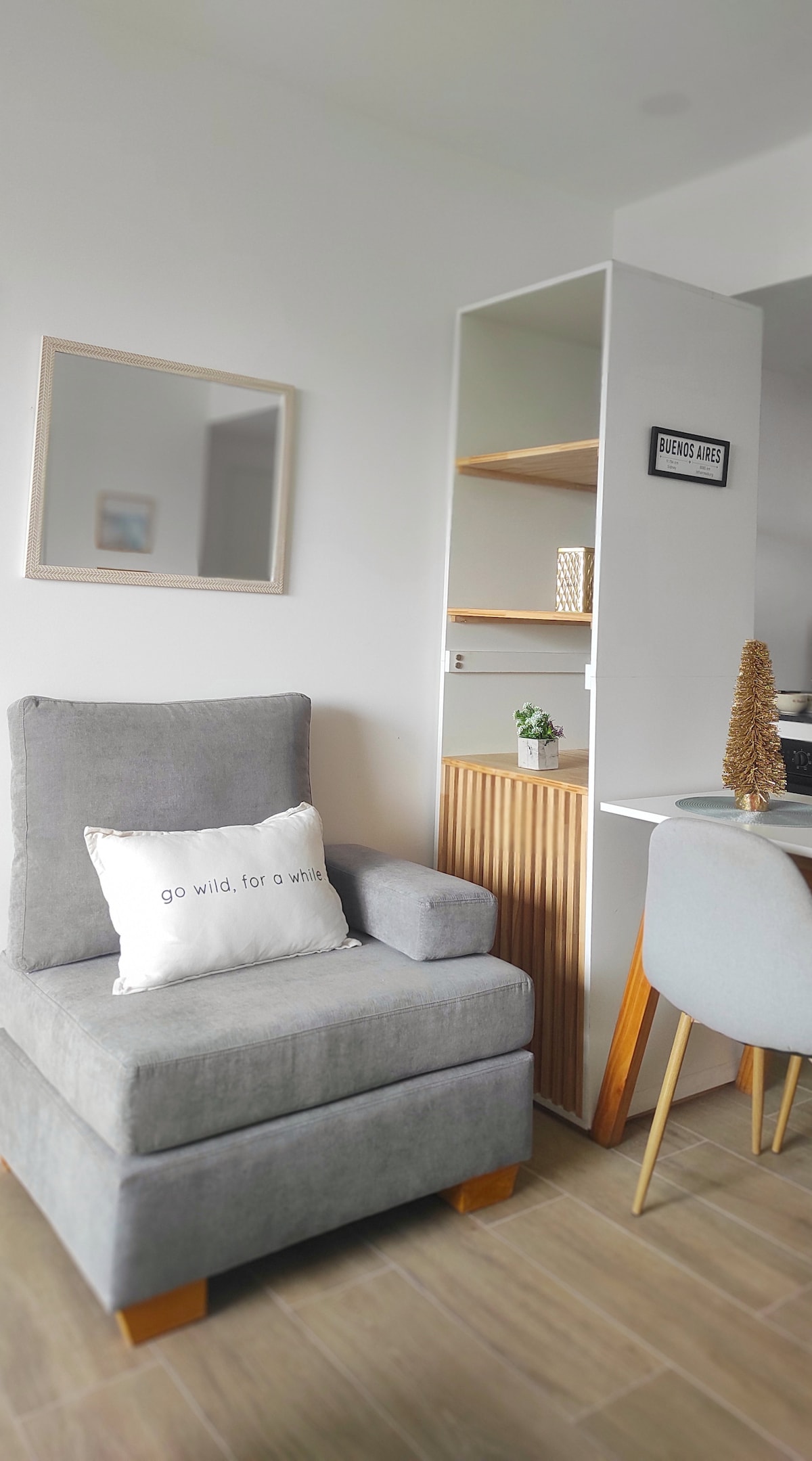 A comfortable gray armchair sits next to a small wooden shelf, showcasing decorative items. A mirror is mounted on the wall, reflecting natural light. A simple dining table and chair are visible in the background, providing a functional and welcoming space.