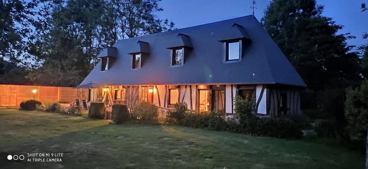 The exterior of a charming cottage is shown in the evening light, highlighting its wooden beams and traditional architecture. Soft lighting from the windows creates a welcoming glow, and the spacious lawn is surrounded by trees and manicured bushes.