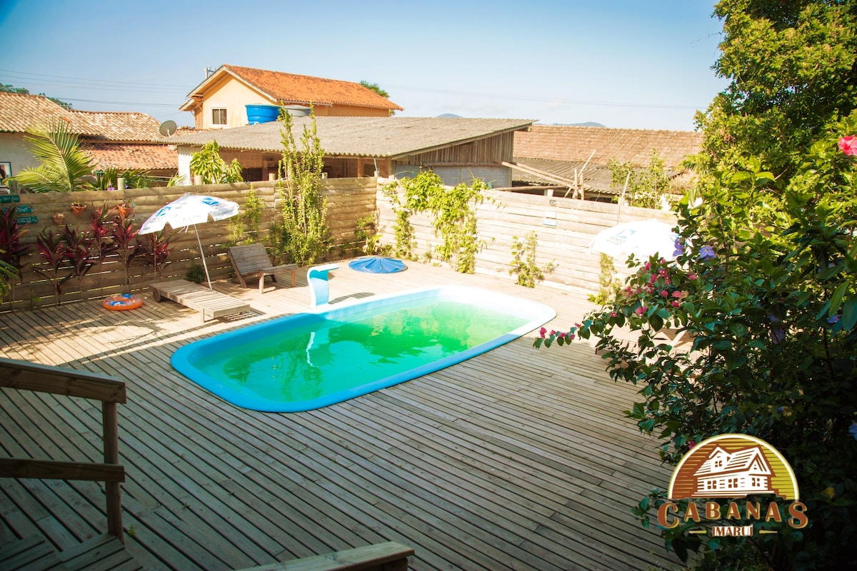 A swimming pool is situated within a wooden deck area, surrounded by lush greenery and colorful plants. Lounge chairs and umbrellas are arranged for relaxation. A sense of tranquility is evident, enhanced by the natural light reflecting off the water.