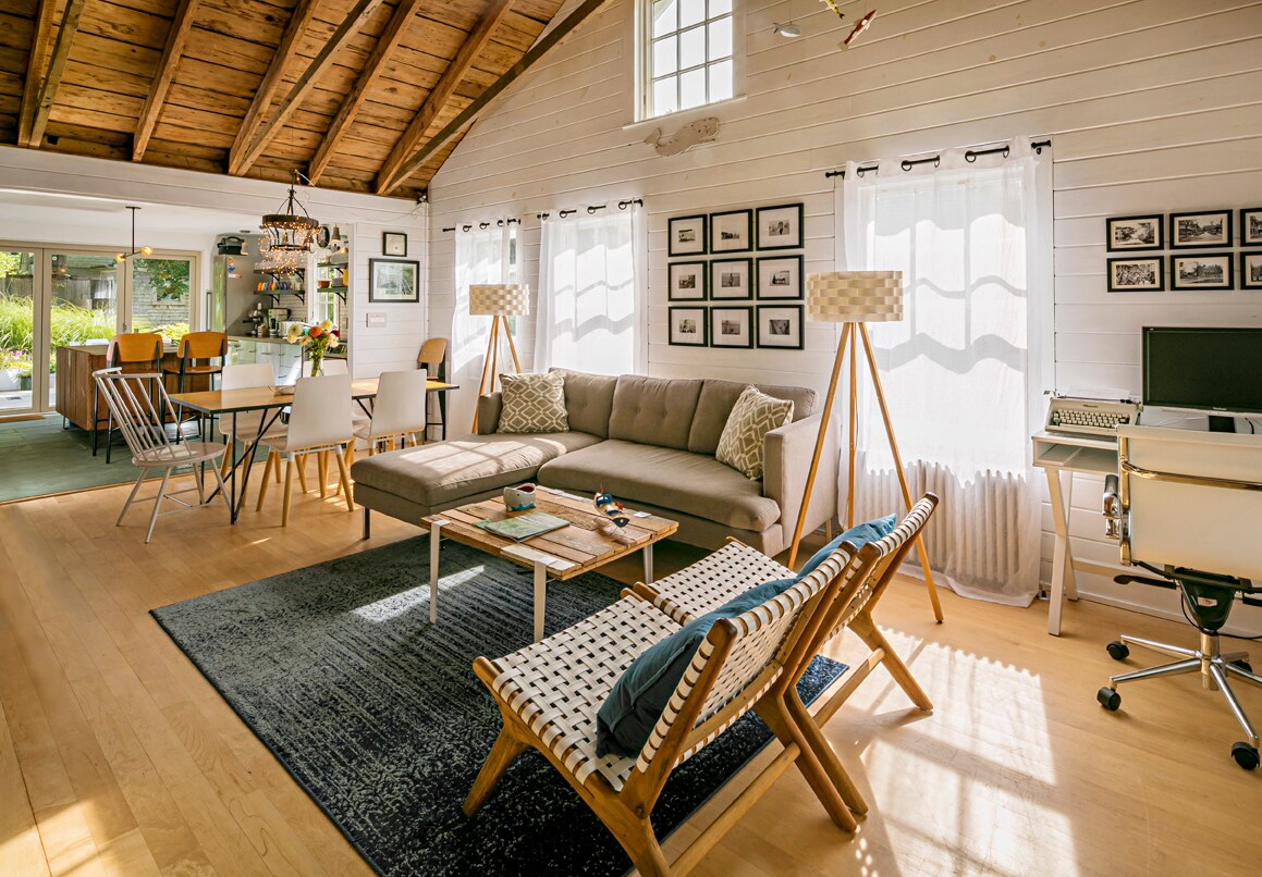 The open living and dining area features a vaulted ceiling with exposed wood beams. A cozy gray sofa is positioned beside two stylish lounge chairs, with a coffee table placed in front. Natural light fills the space through large windows, creating a bright ambiance.