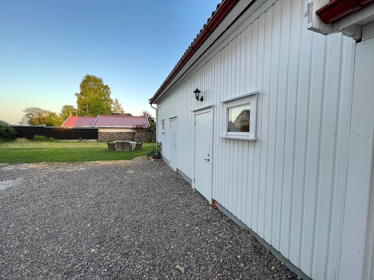 The exterior of the guest annexe features a white wooden facade, with a door and two small windows visible. A gravel area leads to a landscaped garden, where a seating group is available for guests. The setting is surrounded by greenery.