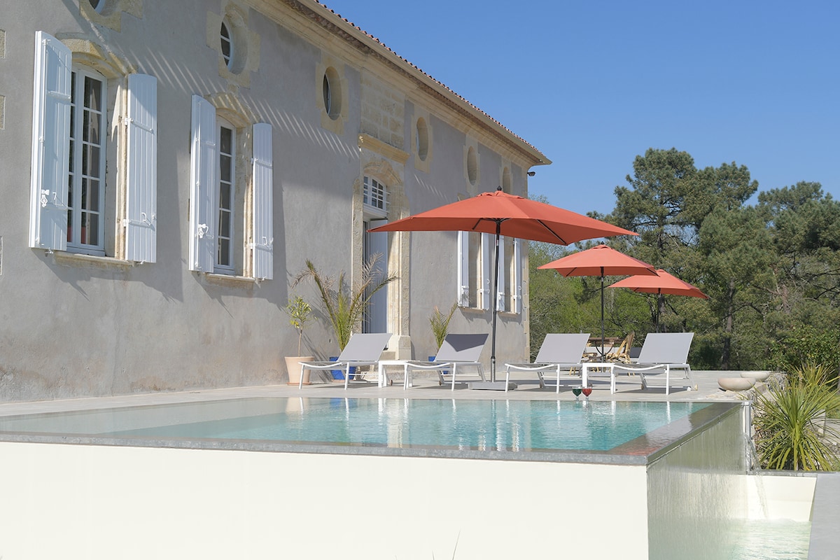 A serene outdoor area is visible, featuring a modern infinity pool with a mirror effect. Multiple sun loungers and umbrellas are arranged beside the pool, against a backdrop of the home's elegant façade and surrounding greenery.