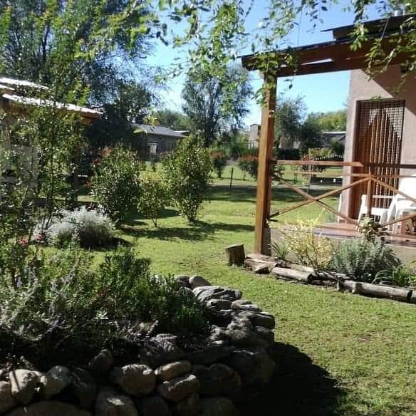 A well-maintained garden is visible, showcasing lush green grass and various plants. A wooden terrace with chairs provides a relaxing spot, while trees frame the space, adding to the serene outdoor environment. Stone borders outline a neatly arranged flower bed.