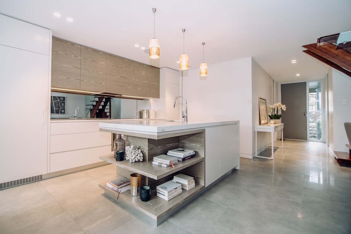A modern kitchen features a large island with open shelving displaying books and decorative items. Sleek cabinetry provides ample storage. Pendant lights hang above, illuminating the space, while a neutral palette enhances the contemporary design. A staircase is visible in the background.