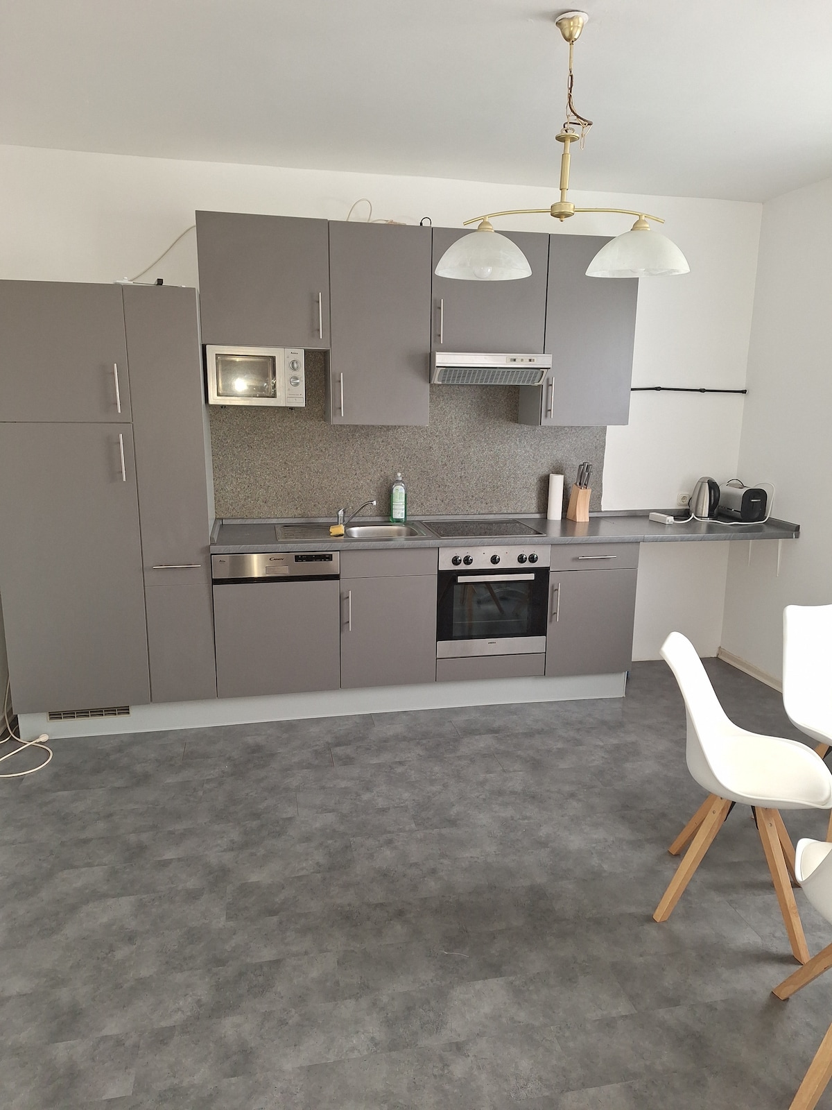 A modern kitchen features grey cabinetry and stainless steel appliances, including an oven and microwave. Counter space is complemented by a neutral backsplash. A small dining table with white chairs is positioned nearby, while pendant lights illuminate the area.