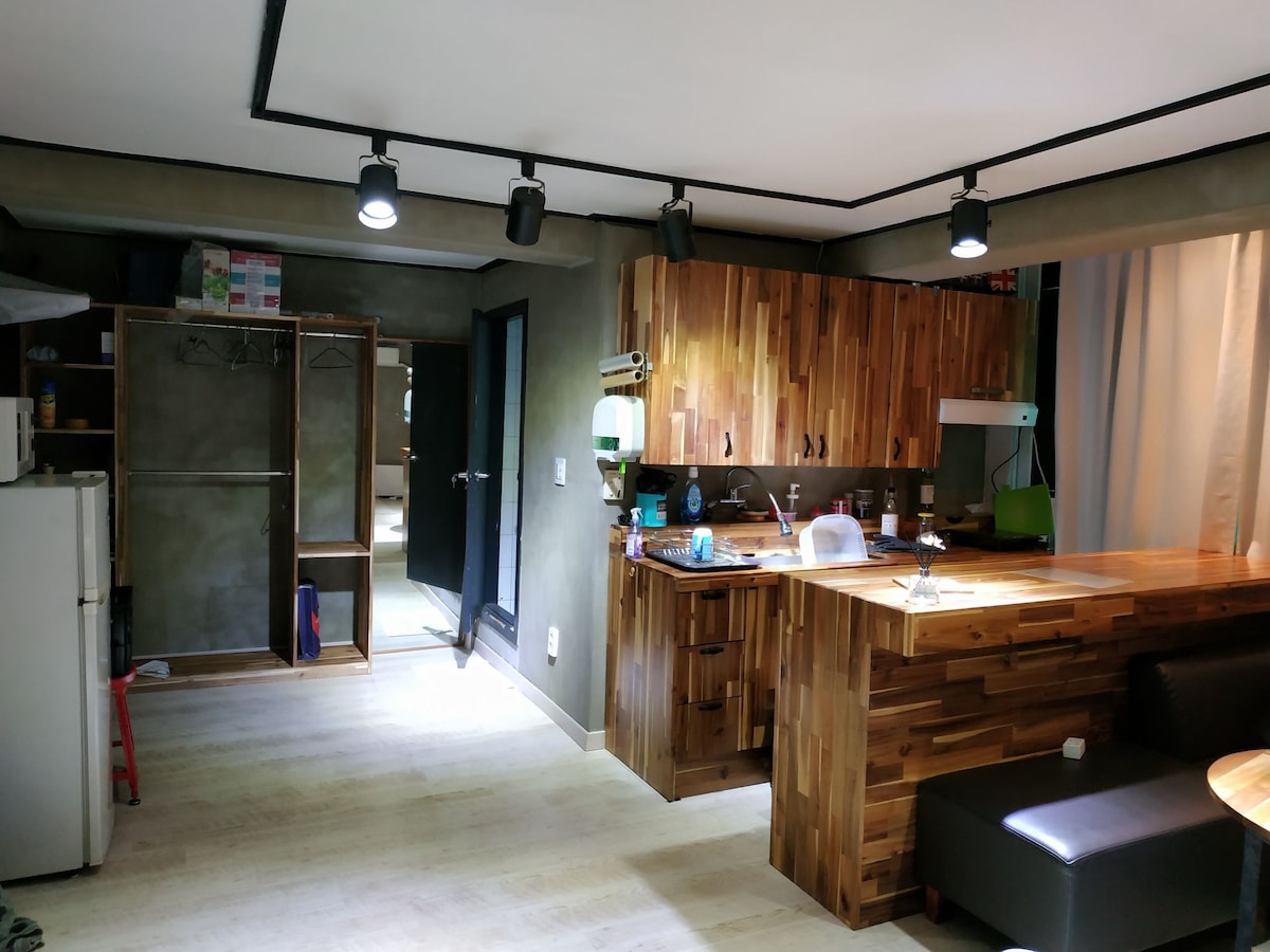 A modern kitchen area features warm wooden cabinetry and a spacious countertop. A dining area with a dark leather seat complements the space. A refrigerator is positioned in the corner, while closet space is visible in the adjacent area, illuminated by overhead track lighting.