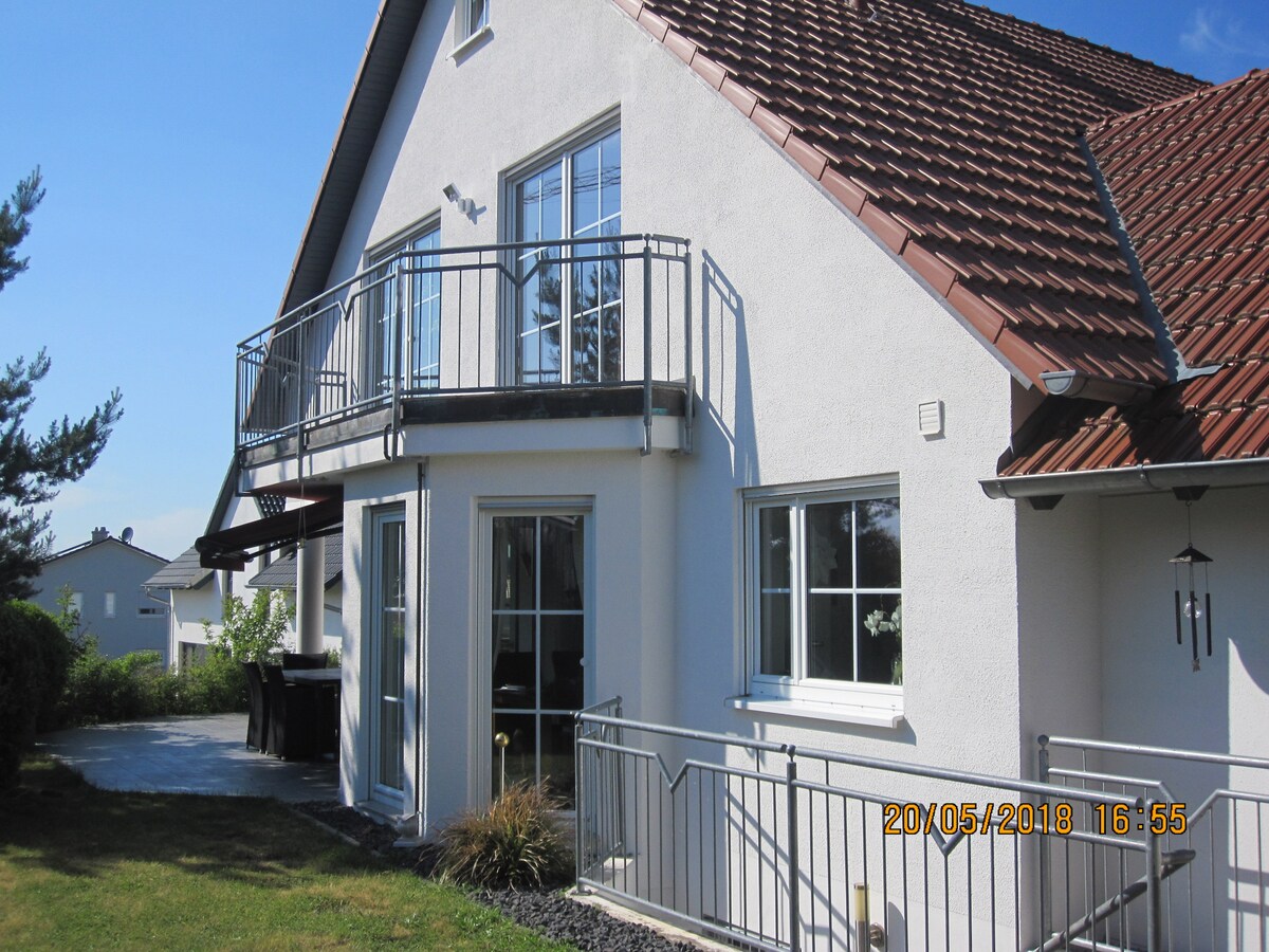 The exterior of the house is shown, featuring a white façade and a sloped, tiled roof. A spacious balcony with a metal railing extends from the upper floor, while large glass doors lead to a patio area surrounded by greenery.