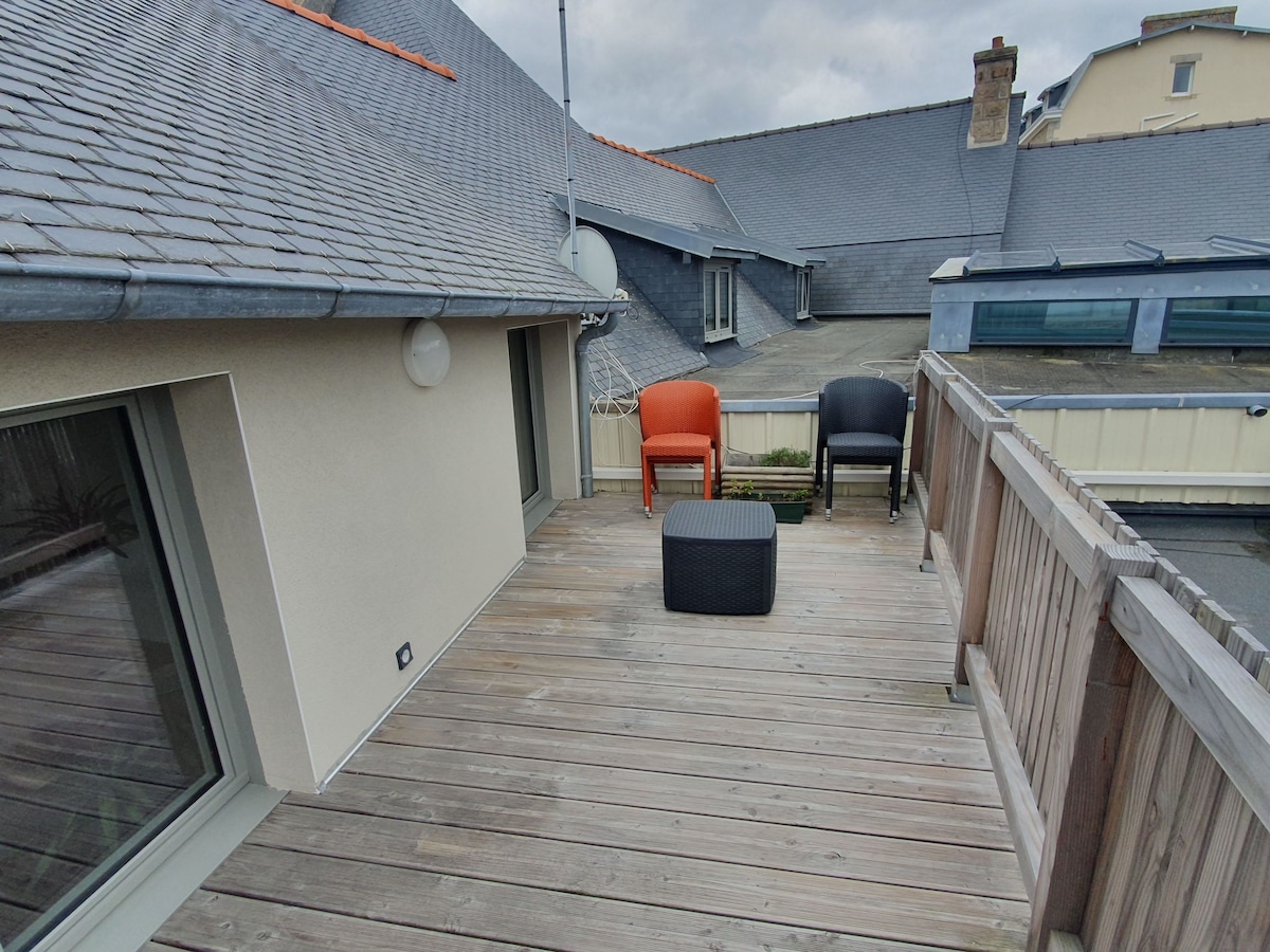 A private terrace features wooden decking, furnished with an orange armchair, a black armchair, and a small black table. Light grey walls are visible, and a sloped gray roof adds to the backdrop. The area offers ample space for relaxation.