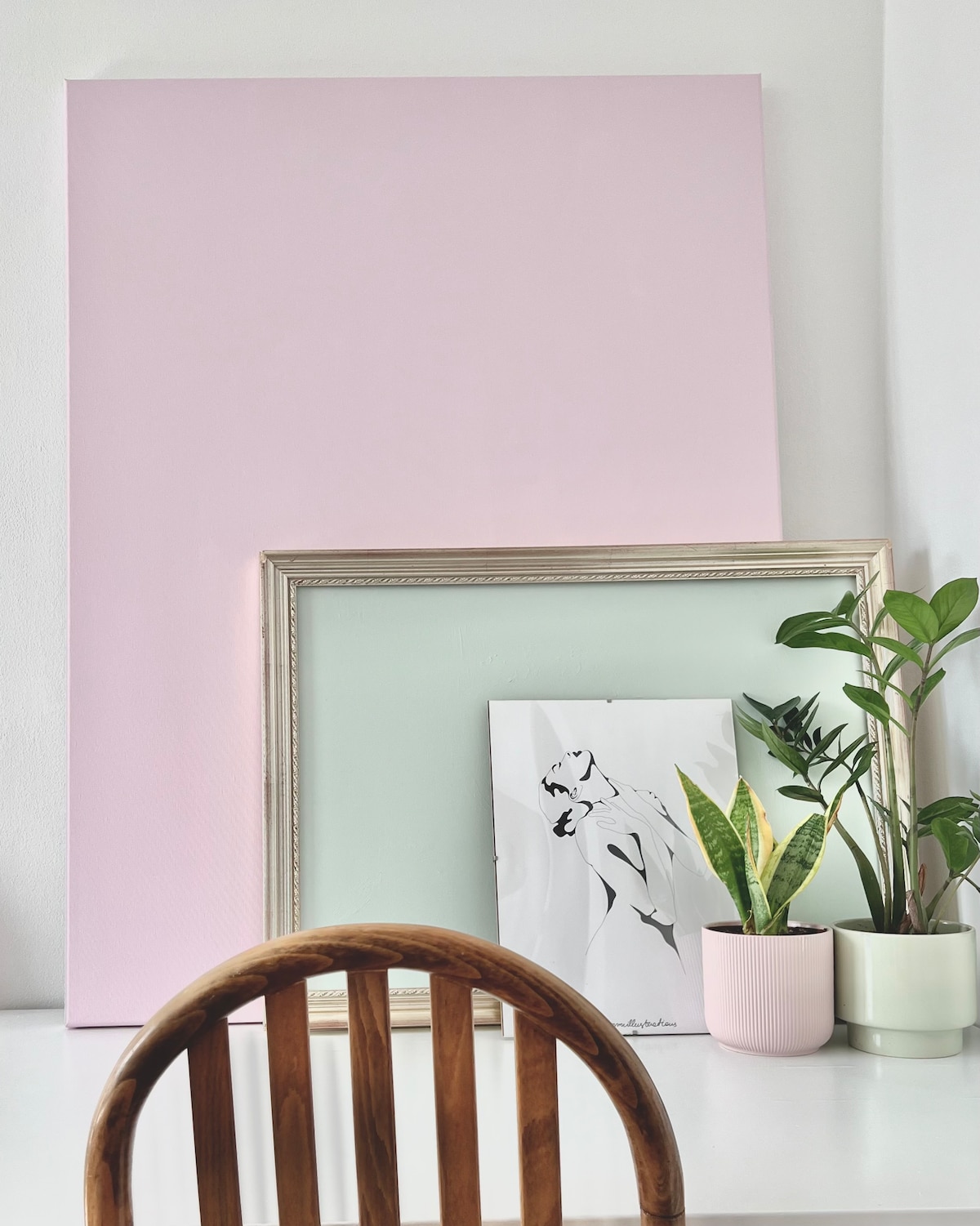 A minimalist display features a light pink canvas leaning against a wall, with a pastel green frame and an art piece showcasing a silhouette. A small potted plant, alongside another green plant, adds a touch of nature to the scene. A wooden chair is positioned nearby.