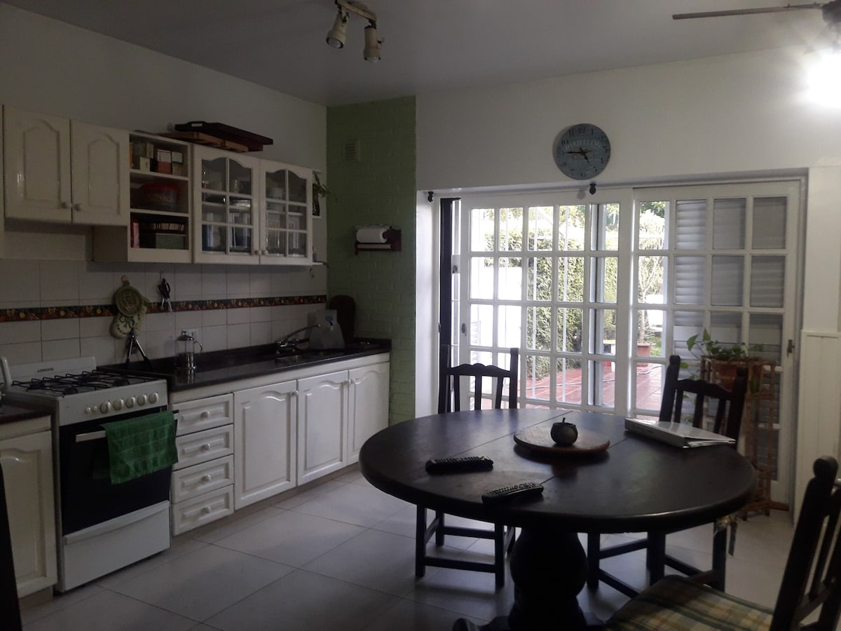A bright kitchen area is visible, featuring white cabinetry and a dark countertop. A round dining table is centrally placed, surrounded by wooden chairs. Natural light filters in through glass doors leading to an outdoor space, with greenery visible beyond.