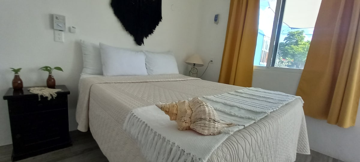 A bright and airy bedroom features a spacious bed with light-colored linens and decorative throw. A bedside table holds potted plants, while a large shell decorates the bed. Natural light enters through a window, complemented by soft yellow curtains.