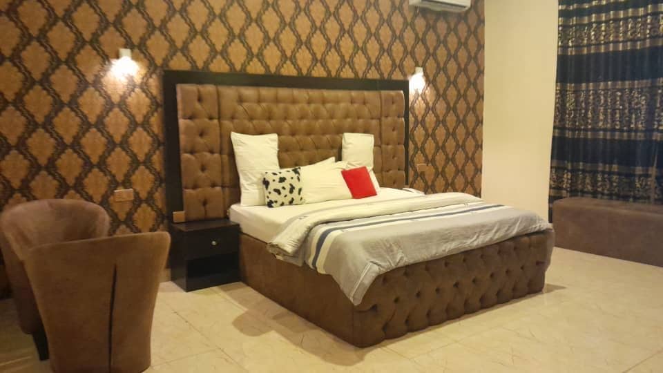 A spacious room features a large upholstered bed with a light-colored blanket and decorative pillows. The accent wall displays an intricate pattern. Two cozy chairs complement the decor, and window treatments add elegance, allowing natural light to filter into the space.