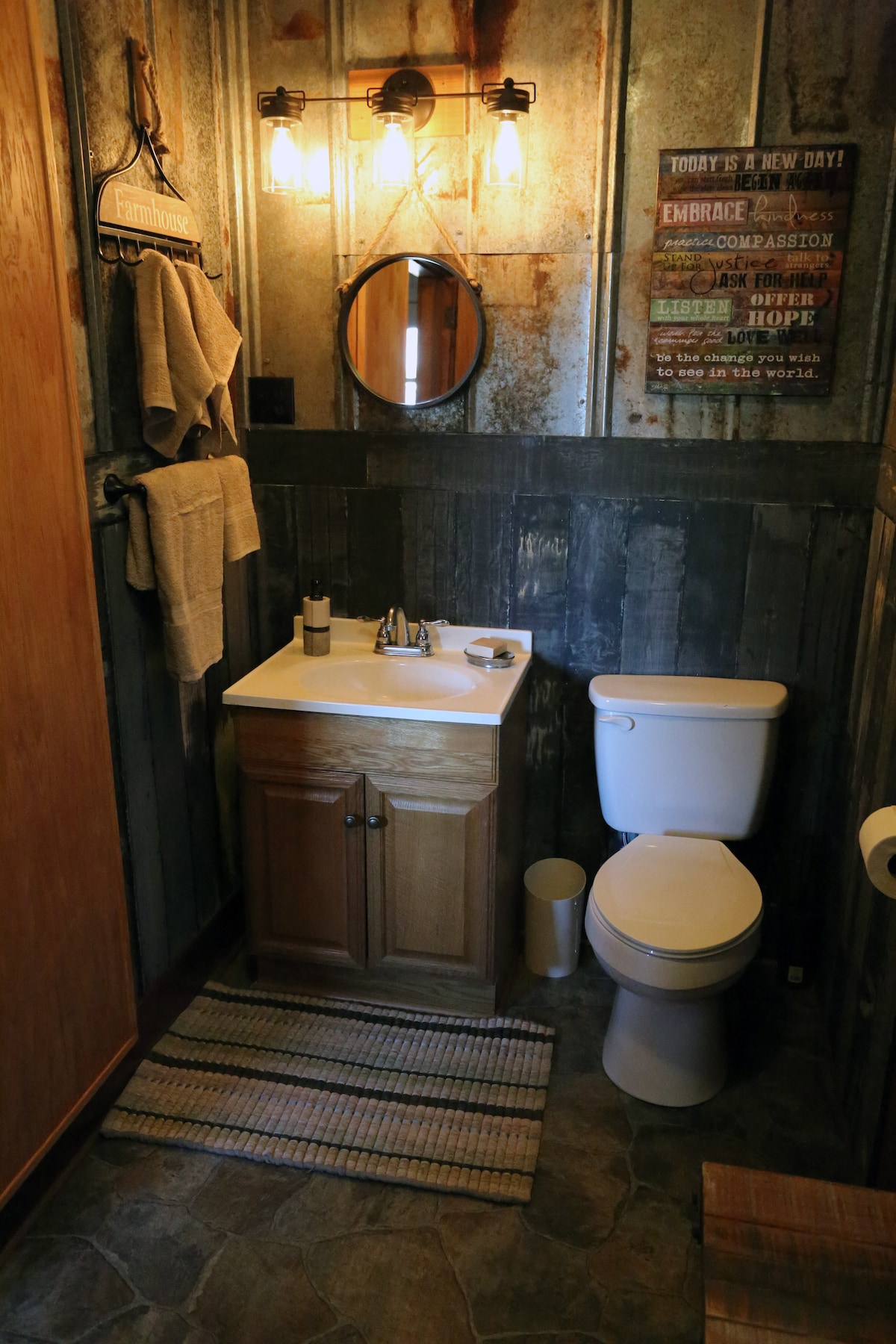 A compact bathroom features a wooden vanity with a sink and storage underneath. A toilet is positioned beside it, with textured walls adding visual interest. Soft lighting is provided by two exposed bulbs above a round mirror. Towels are neatly hung on a hook, enhancing convenience.