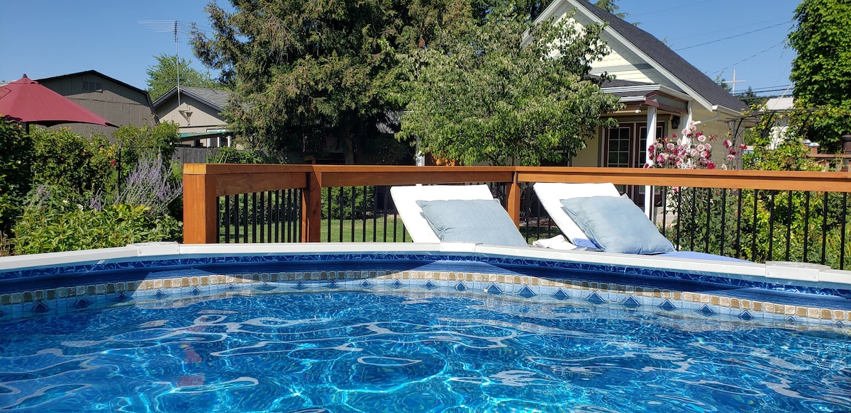 The image captures a seasonal above-ground pool, showcasing refreshing blue water under sunlight. Comfortable lounge chairs with soft cushions are positioned beside the pool, surrounded by lush greenery and flowers. In the background, the cottage and garden area are visible, enhancing the serene atmosphere.
