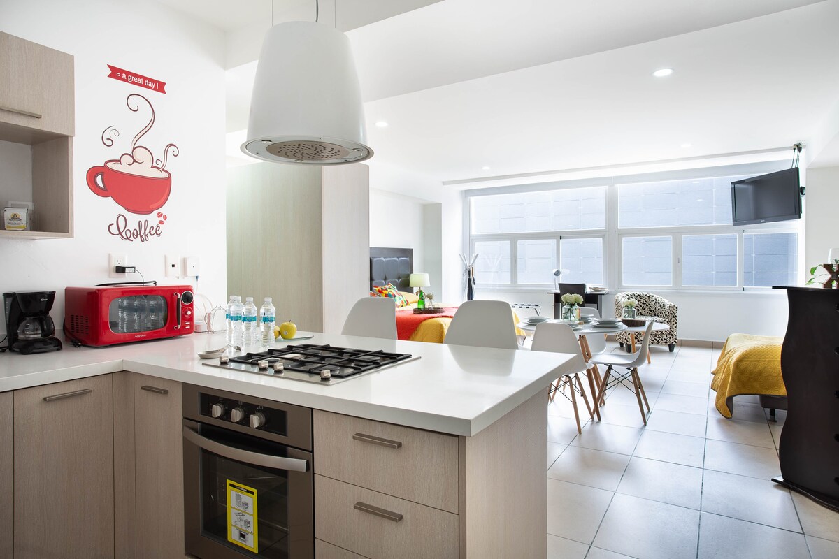 A light and modern kitchen is showcased, featuring a gas stove, microwave, and refrigerator. Bottled water is arranged on the counter. The open layout leads to a dining area with white chairs and a living space adorned with a colorful blanket. Large windows provide ample natural light.