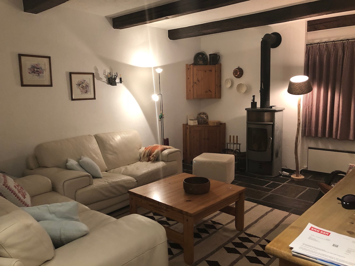A spacious living room features two light-colored sofas and a wooden coffee table. A wood stove stands against a wall, with stacked firewood nearby. Wall art and a lamp add to the cozy ambiance. Large windows are dressed with curtains that allow natural light to filter in.