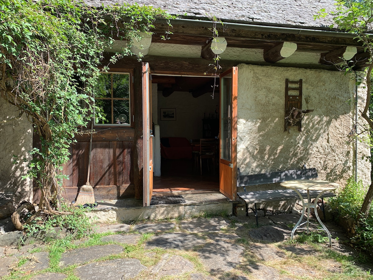 The entrance of the rustic home is framed by lush greenery, including climbing plants along the wall. Wooden doors stand open, revealing a glimpse of the interior. A small table with two chairs is positioned outside on a stone patio, providing a serene outdoor seating area.