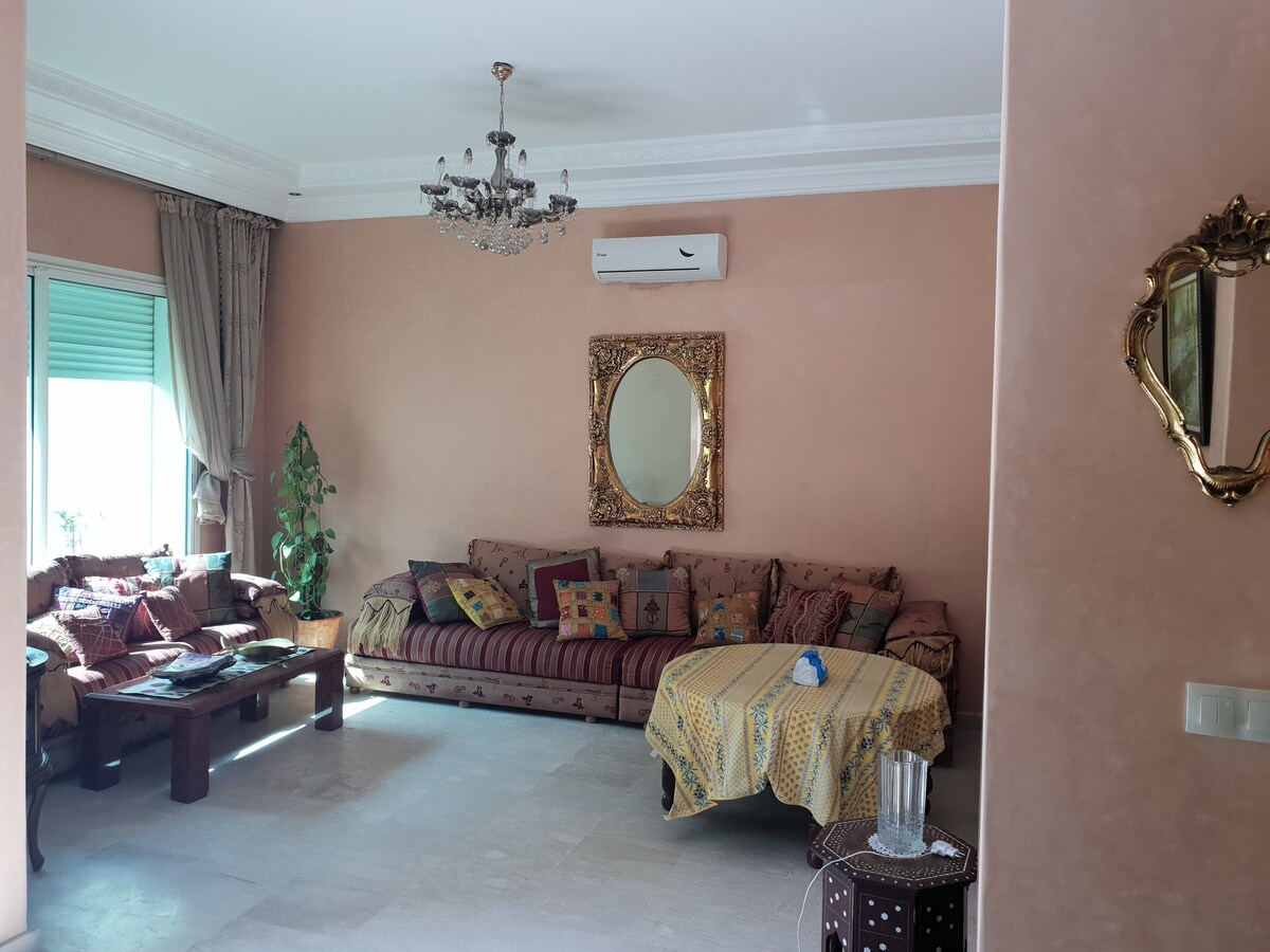 A spacious living area features a plush sofa adorned with decorative cushions and a stylish coffee table. Natural light enters through large windows, complemented by neutral-toned walls and a glamorous chandelier. A decorative mirror adds an elegant touch to the inviting space.