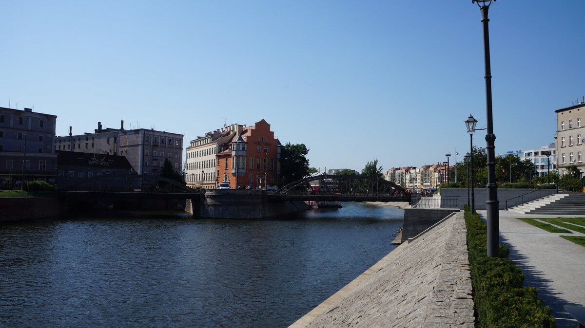 A serene view of a waterfront lined with historic buildings and modern architecture. A pedestrian bridge spans the river, and a promenade featuring landscaped areas and lampposts follows the water's edge, inviting leisurely strolls along the scenic route.