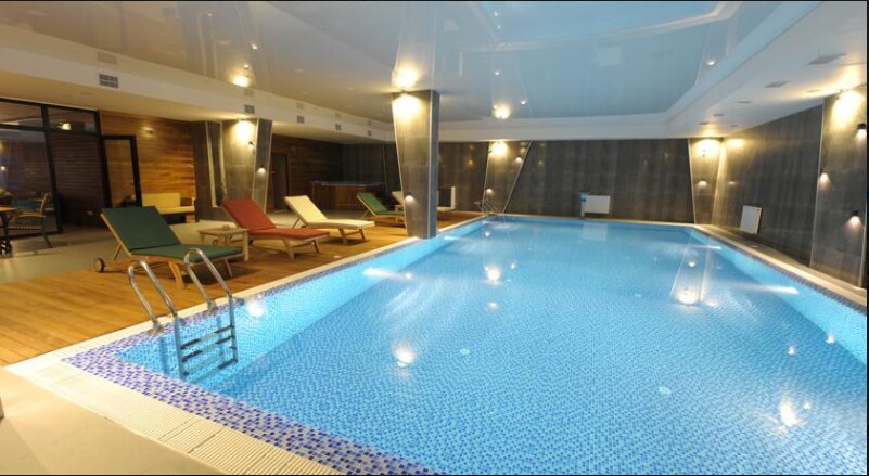 A spacious indoor swimming pool is surrounded by wooden decking. Lounge chairs are arranged on one side, with an inviting turquoise water surface reflecting the ambient lighting. The area is enhanced by modern finishes and subtle wall lighting, creating a calm atmosphere for relaxation.