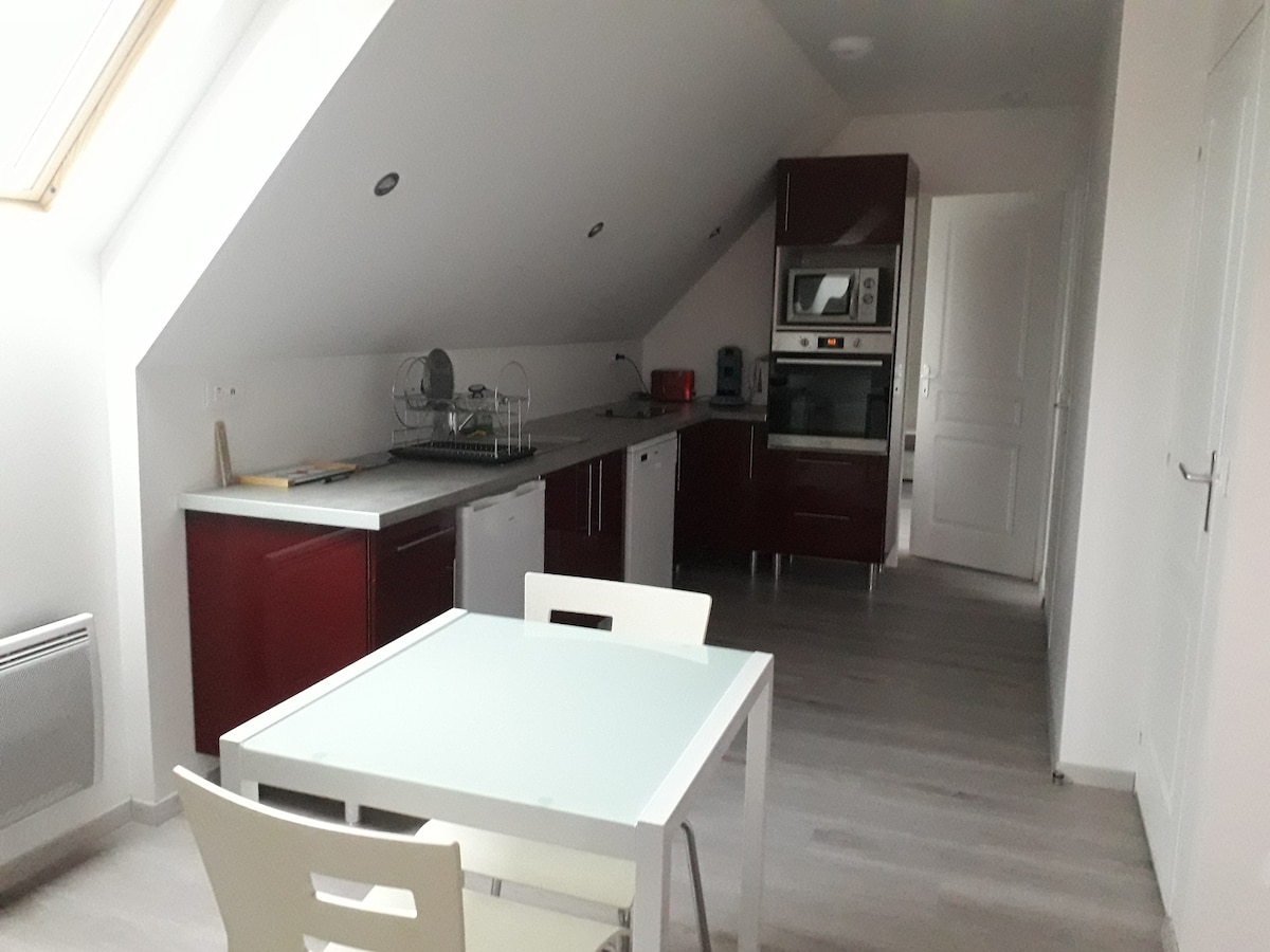 A modern kitchen is presented with sleek red cabinetry and spacious counter surfaces. Essential appliances, including an oven and refrigerator, are clearly visible. A small dining table with white chairs occupies the center, complemented by natural light from the skylight above.