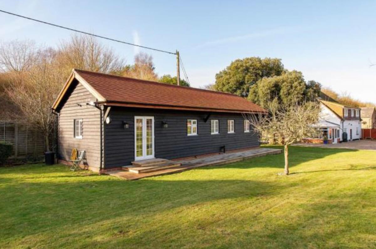 The exterior of a single-story lodge is featured, showcasing a dark wood facade and a sloped roof. A manicured lawn surrounds the structure, with a pathway leading to the entrance. Nearby, trees and another building are visible, creating a serene, rural atmosphere.