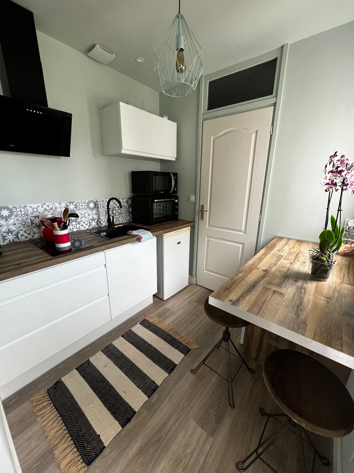 A functional kitchen is depicted, featuring compact cabinetry and modern appliances. A wooden dining table is positioned next to two metal stools. Decorative tile backsplash adds character to the design, while a pendant light provides illumination. Fresh flowers are displayed on the table.