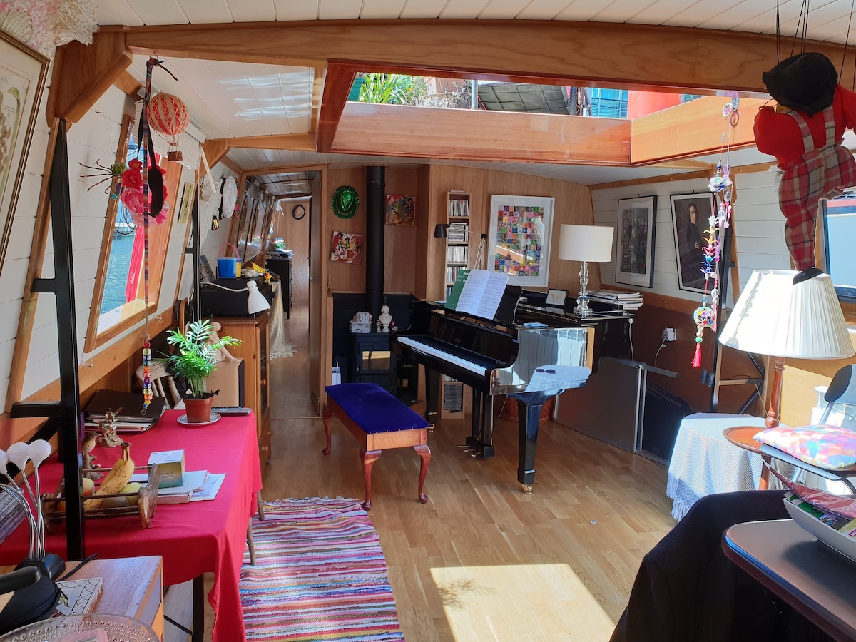 An open-plan living area features warm wooden flooring and ample natural light from skylights. A grand piano is positioned near a colorful rug, surrounded by nautical decor and artwork. A small table with a plant and various items adds to the inviting atmosphere.