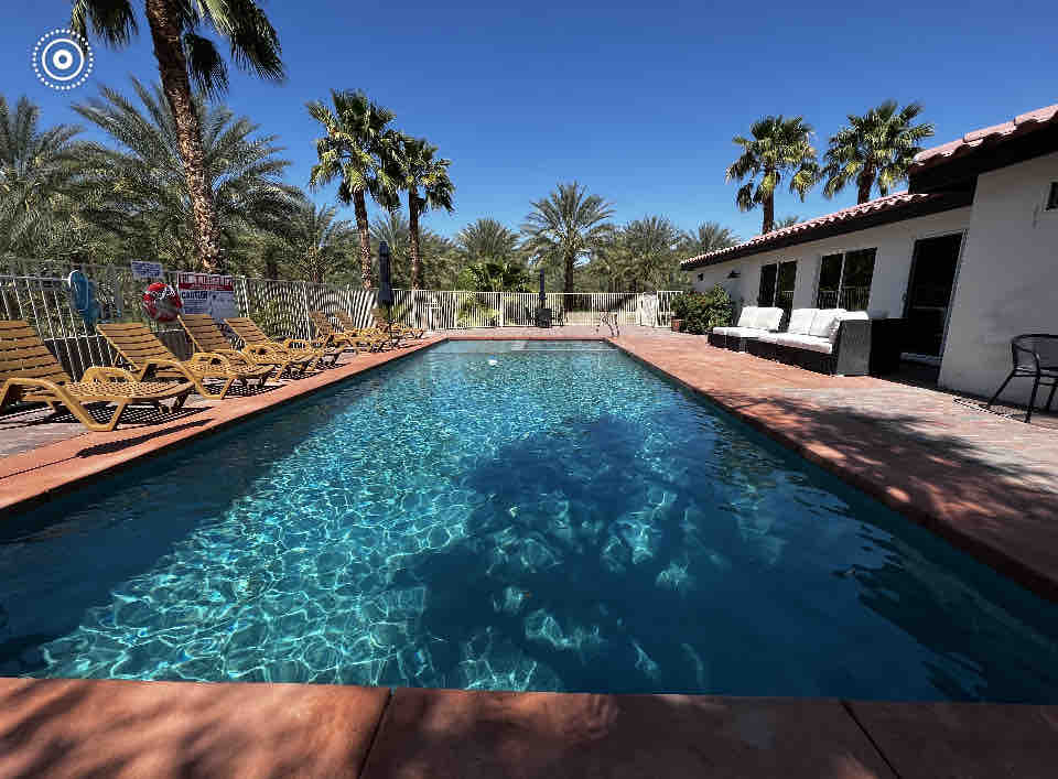 A spacious saltwater pool is surrounded by palm trees, offering a refreshing retreat. Several lounge chairs are arranged along one side, providing plenty of seating options. The area features a clean tiled deck and direct access to the nearby outdoor seating arrangement.