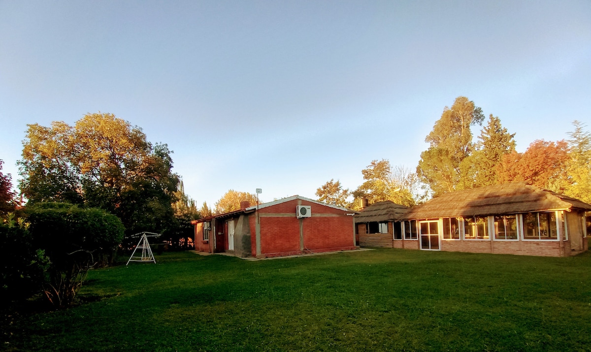 A well-maintained outdoor area is surrounded by trees and greenery. Two distinct buildings are visible, one with a thatched roof and large windows, while the other is finished in red brick. A playground swing set is positioned on the neatly manicured lawn.