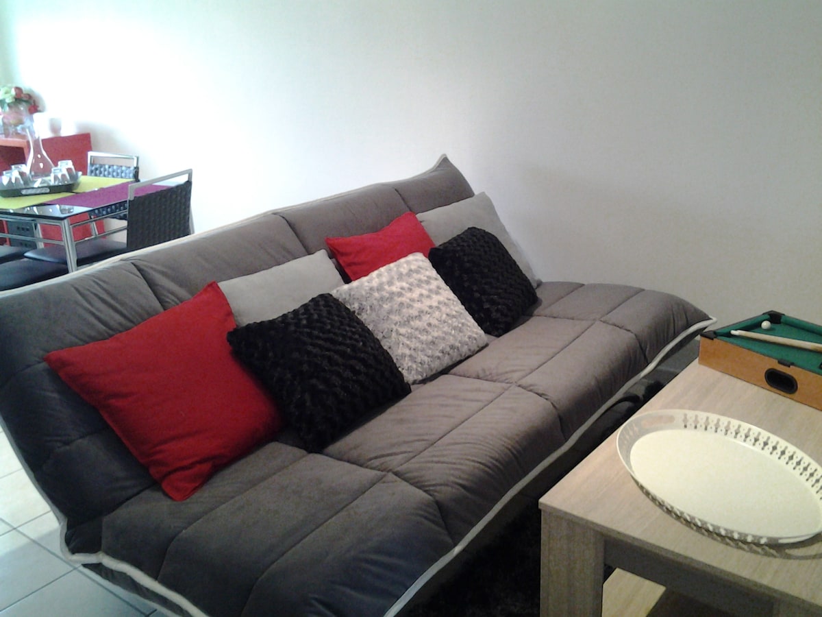 A spacious sofa bed is positioned in the living area, adorned with decorative pillows in various textures and colors. A simple wooden table is nearby, accompanied by a games setup, creating a relaxed atmosphere for leisure activities.