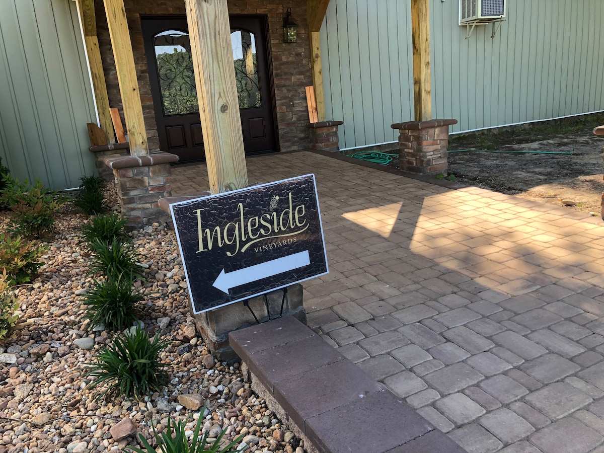 A paver stone walkway leads toward an entrance, framed by wooden pillars. To the left, a sign indicating 'Ingleside Vineyard' points towards the direction of the vineyard, surrounded by neat landscape features and greenery.