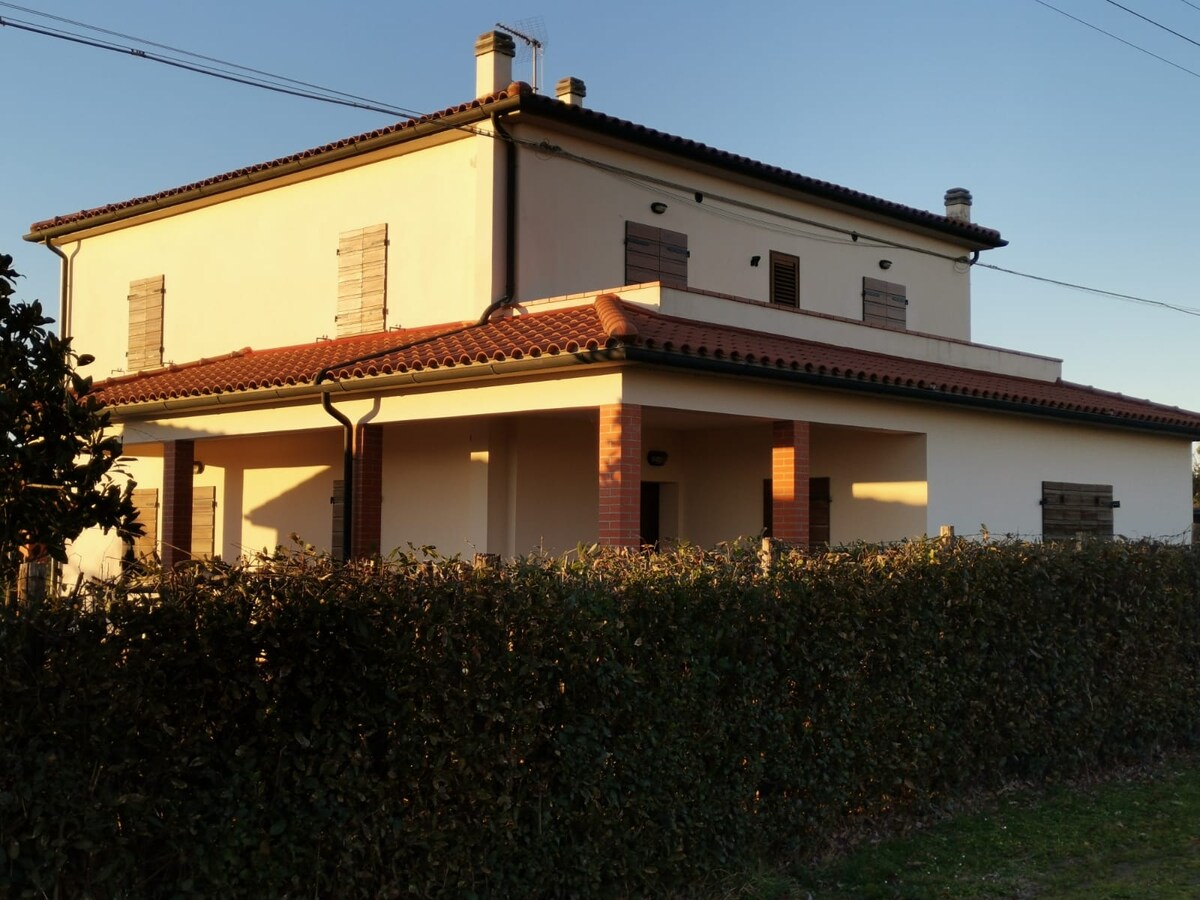 A two-story house with a terracotta roof is surrounded by neatly trimmed hedges. Wooden shutters adorn the windows, and outdoor lighting casts a warm glow on the exterior. The structure features a spacious porch area accessible from the front.