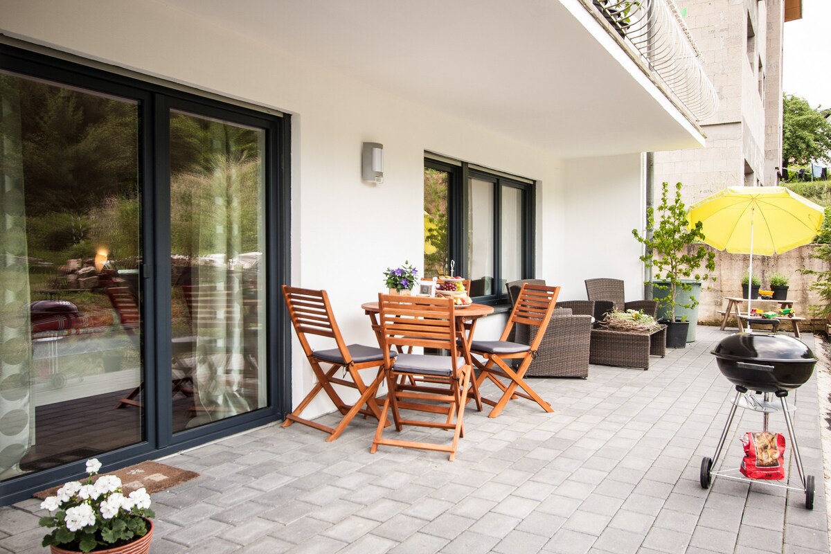 A spacious outdoor patio is furnished with a wooden dining table surrounded by four folding chairs. Comfortable lounge chairs are positioned nearby, and a bright yellow umbrella provides shade. A small grill stands on wheels, and potted plants add greenery to the area.